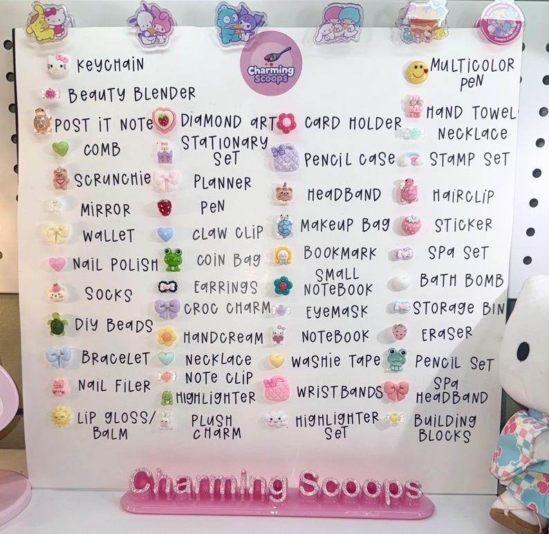 Op de afbeelding: Een wit displaybord met diverse kleine, kleurrijke accessoires, waaronder sleutelhangers, scrunchies en stickers. Het bord heeft handgeschreven labels en de tekst "Charming Scoops."