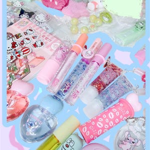 Op de afbeelding: Een verzameling kleurrijke lipgloss, parfums en accessoires. De afbeelding toont verschillende tinten roze, blauwe en transparante lipgloss tubes met glitter en karakterontwerpen. Andere items zijn een parfumfles, een notitieblok en sleutelhangers.