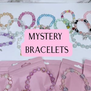May include: A collection of colorful beaded bracelets in various shades of pink, blue, green, and purple. The bracelets are displayed with the text "MYSTERY BRACELETS" on a pink background, and are packaged in pink pouches.