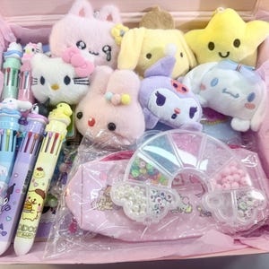 Op de afbeelding: Een roze doos gevuld met pluche speelgoed en briefpapier. De doos bevat pluche dieren van een kat, konijn, ster en andere karakters. Ook inbegrepen zijn multi-color pennen en een roze etui met een regenboogvormige kralenorganizer.