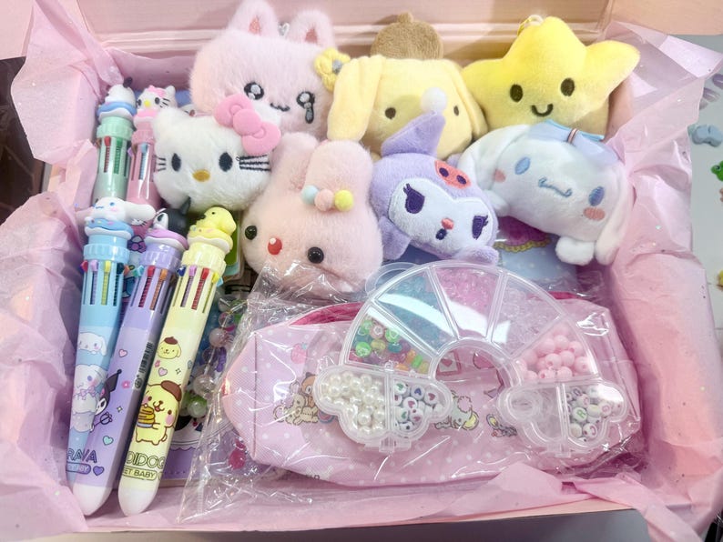 Op de afbeelding: Een roze doos gevuld met pluche speelgoed en briefpapier. De doos bevat pluche dieren van een kat, konijn, ster en andere karakters. Ook inbegrepen zijn multi-color pennen en een roze etui met een regenboogvormige kralenorganizer.