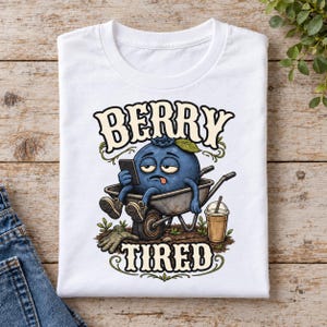 May include: A white t-shirt featuring a graphic of a tired blue blueberry character sitting in a metal wheelbarrow, holding a smartphone, with a coffee cup nearby. The text BERRY TIRED is printed in a bold, decorative font above and below the character.