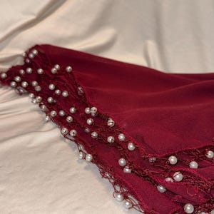 Puede incluir: Bufanda de color rojo oscuro con borde de cuentas. La bufanda está doblada, revelando múltiples capas de tela. Los bordes están adornados con pequeñas cuentas blancas, similares a perlas, que añaden un toque de elegancia. El fondo es de color claro y neutro.