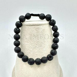 Könnte beinhalten: Armband mit schwarzen Lavasteinperlen. Das Armband besteht aus runden, porösen Perlen, die auf einem schwarzen Band aufgereiht sind. Die Perlen haben eine strukturierte Oberfläche und ein mattes Finish. Das Armband wird auf einer weißen Oberfläche präsentiert.