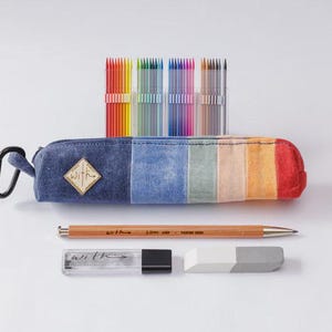 Op de afbeelding: Een denim etui met een regenboogstreepontwerp, een zwarte karabijnhaak en een gouden ruitvormige patch met het woord "WITH". De set bevat kleurpotloden, een houten potlood, een potloodvulling en een gum.