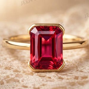 Op de afbeelding: Een gouden ring met een grote, rechthoekige, robijnrode edelsteen. De edelsteen is gezet in een gouden bezel en de band is van effen goud. De ring wordt getoond op een crèmekleurige, kantachtige stof.