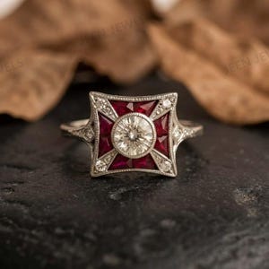 May include: A silver-toned ring featuring a central round brilliant-cut white stone surrounded by four triangular red rubies arranged in a square frame. The metal setting is accented with small white stones along the band and the square edges.