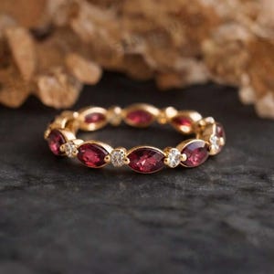 May include: A gold eternity band featuring an alternating pattern of marquise-cut deep red garnet gemstones and small round brilliant-cut clear white diamonds set in a continuous circular arrangement around the entire circumference of the ring.