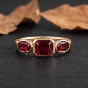 Puede incluir: Un anillo de oro con una piedra preciosa roja central de corte esmeralda, flanqueada por dos piedras rojas más pequeñas de corte ovalado. Las piedras están engastadas en un bisel sobre una banda de oro lisa, creando un diseño de tres piedras.