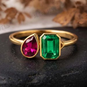May include: A gold ring featuring two prominent gemstones: a pear-shaped deep pink stone and a rectangular emerald-cut vibrant green stone. The stones are set side-by-side on a polished gold band, creating a distinct two-stone design.
