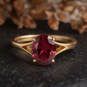 Puede incluir: Un anillo de oro con una gran piedra preciosa de color rojo intenso y corte ovalado, engastada en cuatro garras. La banda presenta un diseño de brazo dividido que se estrecha hacia la piedra central, creando un aspecto pulido y refinado.