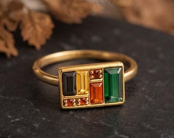 May include: A gold ring featuring a rectangular face set with a collection of multi-coloured gemstones. The arrangement includes a black baguette, a yellow baguette, a green baguette, an orange baguette, and several small round orange stones.