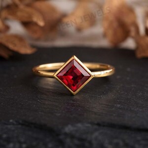 May include: A gold ring featuring a single princess-cut red gemstone set in a square bezel. The band is thin and polished, showcasing a smooth metallic finish. The vibrant red stone is positioned centrally, creating a geometric focal point.