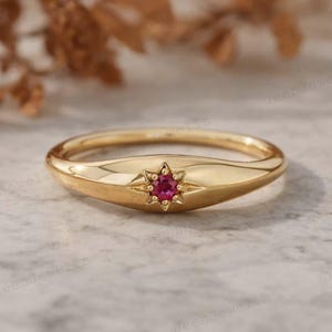 May include: A gold ring with a starburst design featuring a small, round, red gemstone at the center. The ring has a smooth, polished band and a slightly raised setting for the gemstone. The ring is displayed on a light-colored surface.
