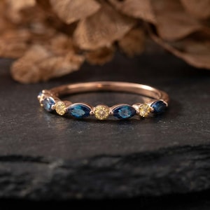 Könnte beinhalten: Ein Ring aus Roségold mit einem abwechselnden Muster aus blauen Steinen im Marquise-Schliff und gelben Steinen im Rundschliff, die entlang der oberen Hälfte des Metallbandes gefasst sind. Der Ring hat eine polierte Oberfläche und ein schmales Profil.