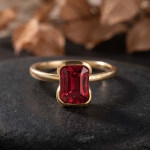 May include: A gold ring featuring a single rectangular emerald-cut red gemstone set in a smooth bezel setting. The band is thin and polished, showcasing a metallic yellow finish that complements the deep red color of the central stone.