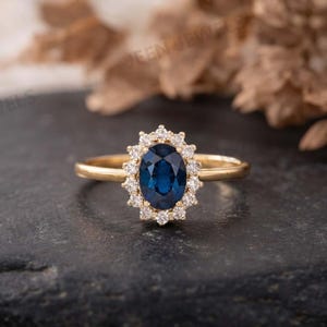 Könnte beinhalten: Ein Goldring mit einem zentralen, oval geschliffenen, tiefblauen Saphir, der von einem Kranz aus kleinen, runden weißen Diamanten umgeben ist. Die Ringschiene ist schmal und aus poliertem Gold gefertigt, was einen klassischen Look erzeugt.