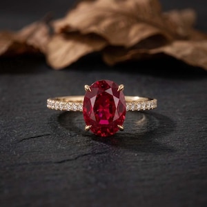 Könnte beinhalten: Ein Goldring mit einem großen, oval geschliffenen, tiefroten Edelstein als Herzstück. Die Ringschiene besteht aus Gelbgold und ist mit einer Reihe kleiner, klarer, rund geschliffener Diamanten besetzt, die beidseitig des roten Steins liegen.