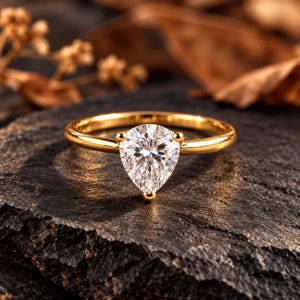 May include: A gold ring featuring a single pear-cut clear gemstone set in a three-prong mounting. The band is smooth and polished, showcasing a reflective metallic finish that complements the faceted stone at the centre of the jewellery piece.