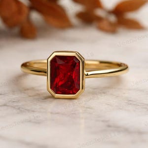 Op de afbeelding: Een gouden ring met een rechthoekige, dieprode edelsteen. De edelsteen is gezet in een gouden bezel, en de band is effen, gepolijst goud. De ring wordt getoond op een lichtgekleurd oppervlak.