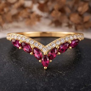 May include: A gold V-shaped ring featuring a row of small round white stones along the top edge and a series of marquise-cut deep red stones set along the bottom curve, creating a chevron design that points downward in the center of the band.
