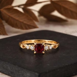 Op de afbeelding: Een gouden ring met een centraal vierkant geslepen dieprode edelsteen, geflankeerd door twee kleine ronde heldere stenen aan weerszijden. De ringband heeft een verfijnd, getextureerd metaalontwerp dat uitloopt vanaf de stenen voor een symmetrisch uiterlijk.