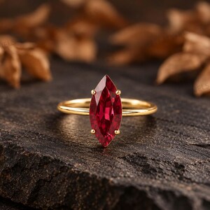 Op de afbeelding: Een gouden ring met een enkele dieprode edelsteen in marquise-slijpvorm, gezet in een zetting met vier pootjes. De ringband is dun en gepolijst en toont een levendige, gefacetteerde rode steen die aan beide uiteinden in een punt uitloopt.