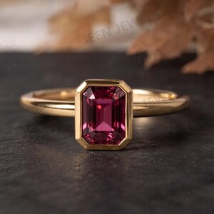 Può includere: Un anello in oro con una pietra preziosa rettangolare color rosa intenso dal taglio smeraldo, montata in una lunetta liscia. La fascia è in oro lucido con un design semplice e sottile che mette in risalto la pietra centrale e il colore.
