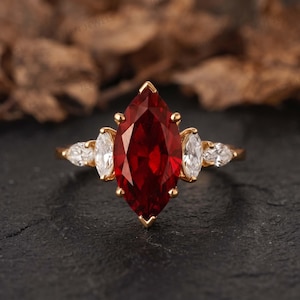 Peut inclure: Une bague en or avec une grande pierre précieuse rouge foncé taille marquise au centre, flanquée de deux pierres blanches transparentes taille marquise de chaque côté, montée sur un anneau en or jaune poli avec un sertissage à griffes.