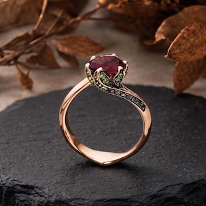 Op de afbeelding: Een roségouden ring met een grote, ronde, dieprode middensteen. De zetting heeft de vorm van een bloemknop met kleine groene accentstenen die de bloemblaadjes vormen en een rij kleine zwarte steentjes langs de band van de ring.