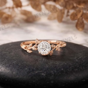 May include: A rose gold ring featuring a central round brilliant-cut white gemstone. The band is designed with delicate metal leaf motifs extending from the center stone, accented with small embedded white stones along the vine-like structure.