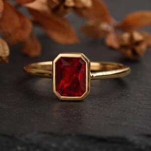 Könnte beinhalten: Ein goldener Ring mit einem rechteckigen, facettierten tiefroten Edelstein in einer glatten Zargenfassung. Die Ringschiene ist schmal und poliert und besticht durch ein klares Design, das die leuchtende Farbe und Reinheit des zentralen Steins betont.