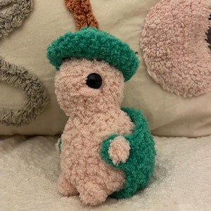 May include: A handmade plush turtle toy with a pink body, a green shell, and a green hat. The turtle has a black eye and is made of a soft, textured material. The toy is on a white surface.