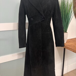 Peut inclure: Un long manteau en daim noir doté d'un large col à revers cranté, de manches longues et d'une taille ajustée avec des surpiqûres horizontales et deux boutons noirs. Le manteau descend jusqu'au milieu du mollet avec un ourlet droit.