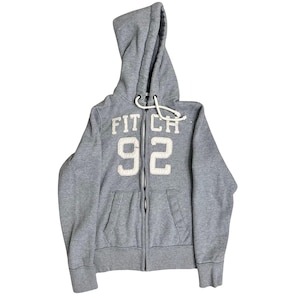 May include: A grey zip-up hoodie with a hood and front pockets. The hoodie features the text "FIT CH 92" in white, stitched lettering. The drawstring is white. The hoodie is made of a soft, comfortable material.