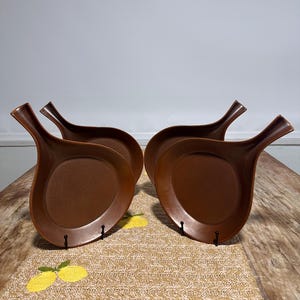 May include: Four brown, handled plates are arranged on a wooden table. The plates have a unique shape, with a rounded bowl and a long, curved handle. The table is covered with a tan runner with yellow lemon designs.