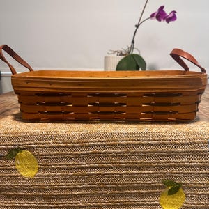 Puede incluir: Una cesta rectangular tejida con un acabado de madera marrón claro y tiras decorativas de color marrón oscuro. La cesta cuenta con dos asas de cuero marrón unidas a los lados. La estructura consiste en tiras de madera tejidas.
