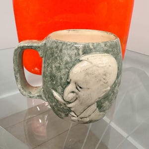 May include: A vintage ceramic mug with a handle, featuring a relief of a man's face. The mug has a mottled green and white glaze, with the face in a lighter color. An orange vase is in the background.