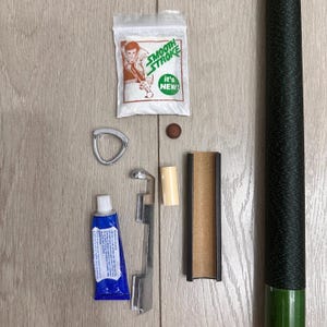 May include: A billiard cue repair kit including a packet of Smooth Stroke powder, a small brown tip, a silver triangular clamp, a blue tube of adhesive, a cylindrical ferrule, a sanding tool, and a green cue shaft with a black textured grip.