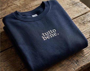 以下が含まれることがあります： 胸の中央に白い刺繍で「tutto bene.」と施された、ネイビーブルーのクルーネックトレーナー。長袖でリブ編みの襟が付いており、ネックラインからは内側のフリース素材が見える状態で折りたたまれています。