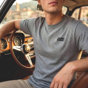 May include: A heather gray short-sleeved t-shirt featuring the text "tutto bene." printed in black on the left chest. The shirt has a crew neckline and a standard fit, shown in a setting with a classic car steering wheel and dashboard.