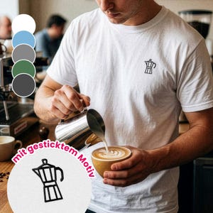 Può includere: T-shirt bianco con un piccolo disegno di caffettiera nera ricamata. L'immagine mostra una persona che versa latte in una tazza di caffè, creando latte art. La maglietta fa parte di una collezione di abbigliamento a tema caffè.