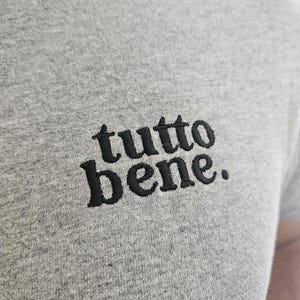 May include: A heather gray t-shirt featuring the text "tutto bene." embroidered in black thread on the chest. The fabric has a visible textured knit pattern, and the lettering is rendered in a bold, lowercase serif font with a period at the end.