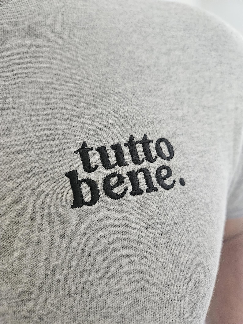 May include: A heather gray t-shirt featuring the text "tutto bene." embroidered in black thread on the chest. The fabric has a visible textured knit pattern, and the lettering is rendered in a bold, lowercase serif font with a period at the end.