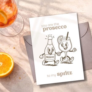 May include: Greeting card with the text "You are the prosecco to my spritz." The card features cartoon illustrations of a prosecco bottle and a cocktail glass holding hands. An orange slice and a cocktail are visible.