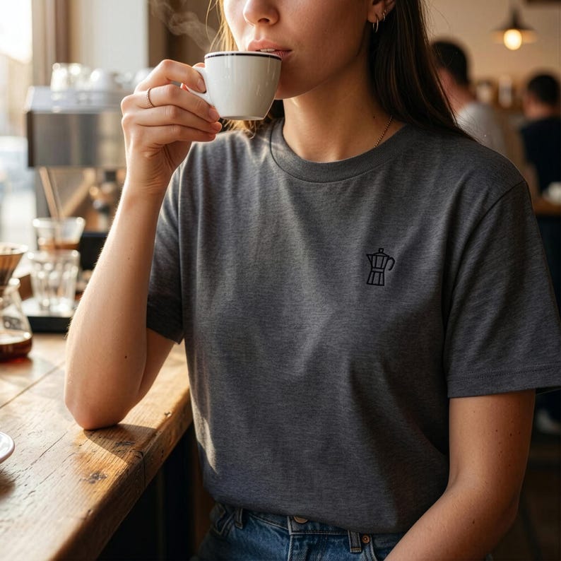 以下が含まれることがあります： 左胸にモカポットの黒い刺繍が施された、ダークグレーの半袖Tシャツ。霜降り調の生地で作られており、ブルーのデニムジーンズと組み合わされています。