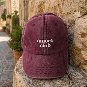 Può includere: Un cappellino da baseball color marrone scuro con struttura a sei pannelli e visiera curva. La parte anteriore presenta la scritta "amore club" ricamata in filo bianco. Il tessuto ha un aspetto slavato con cuciture ben visibili.