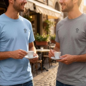以下が含まれることがあります： ライトブルーのTシャツとグレーの杢Tシャツ。どちらも左胸にモカポットの小さな黒い線画がプリントされています。両方のシャツは半袖でクルーネックのデザインであり、無地の生地で作られています。