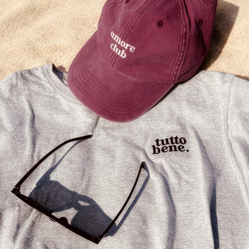 May include: A maroon baseball cap with the white embroidered text "amore club" and a grey sweatshirt featuring the black embroidered text "tutto bene." along with a pair of black-framed sunglasses resting on the fabric surface.