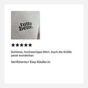 May include: A heather gray t-shirt featuring the black embroidered text "tutto bene." on the chest. The fabric has a visible knit texture, and the lettering is displayed in a lowercase, rounded serif font with a period at the end.